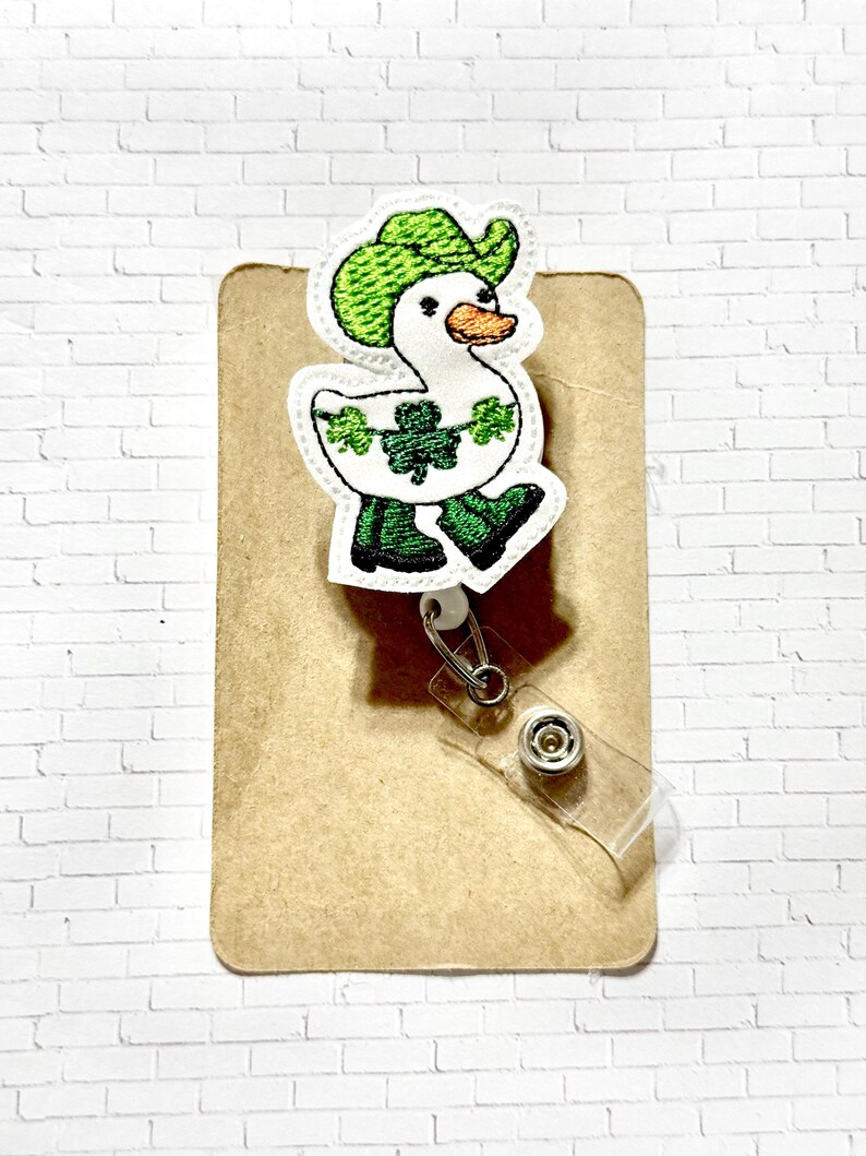 May include: A white goose wearing a green cowboy hat and green boots with shamrocks around its neck. The goose is attached to a retractable badge holder.