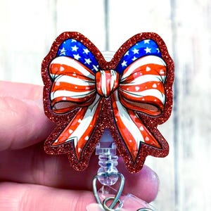 May include: A red glitter badge reel with a bow design. The bow features a patriotic design with red and white stripes, blue stars, and a red glitter outline. The badge reel has a clear plastic clip and a retractable cord.