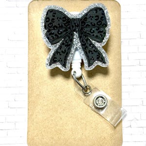 Puede incluir: Un carrete de insignia en forma de lazo negro con un borde de purpurina plateada y un estampado de leopardo negro. El carrete tiene un cord&oacute;n retr&aacute;ctil blanco y una correa de pl&aacute;stico transparente con cierre a presi&oacute;n.