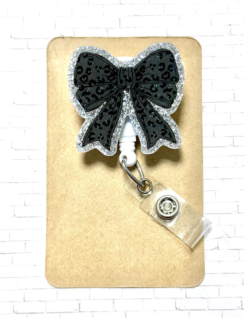 Puede incluir: Un carrete de insignia en forma de lazo negro con un borde de purpurina plateada y un estampado de leopardo negro. El carrete tiene un cord&oacute;n retr&aacute;ctil blanco y una correa de pl&aacute;stico transparente con cierre a presi&oacute;n.