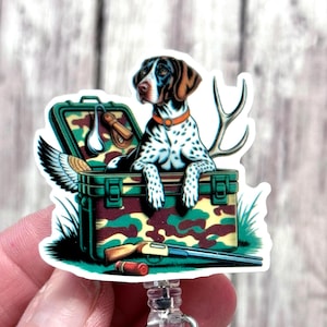 May include: A badge reel featuring a hunting dog with a brown and black head and white spotted body, sitting in a green camouflage box with hunting gear. The box includes a shotgun, duck wing, and antlers.