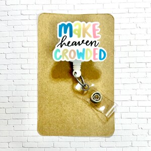 May include: White retractable badge holder with a colorful "Make heaven crowded" graphic. The holder is on a brown background.