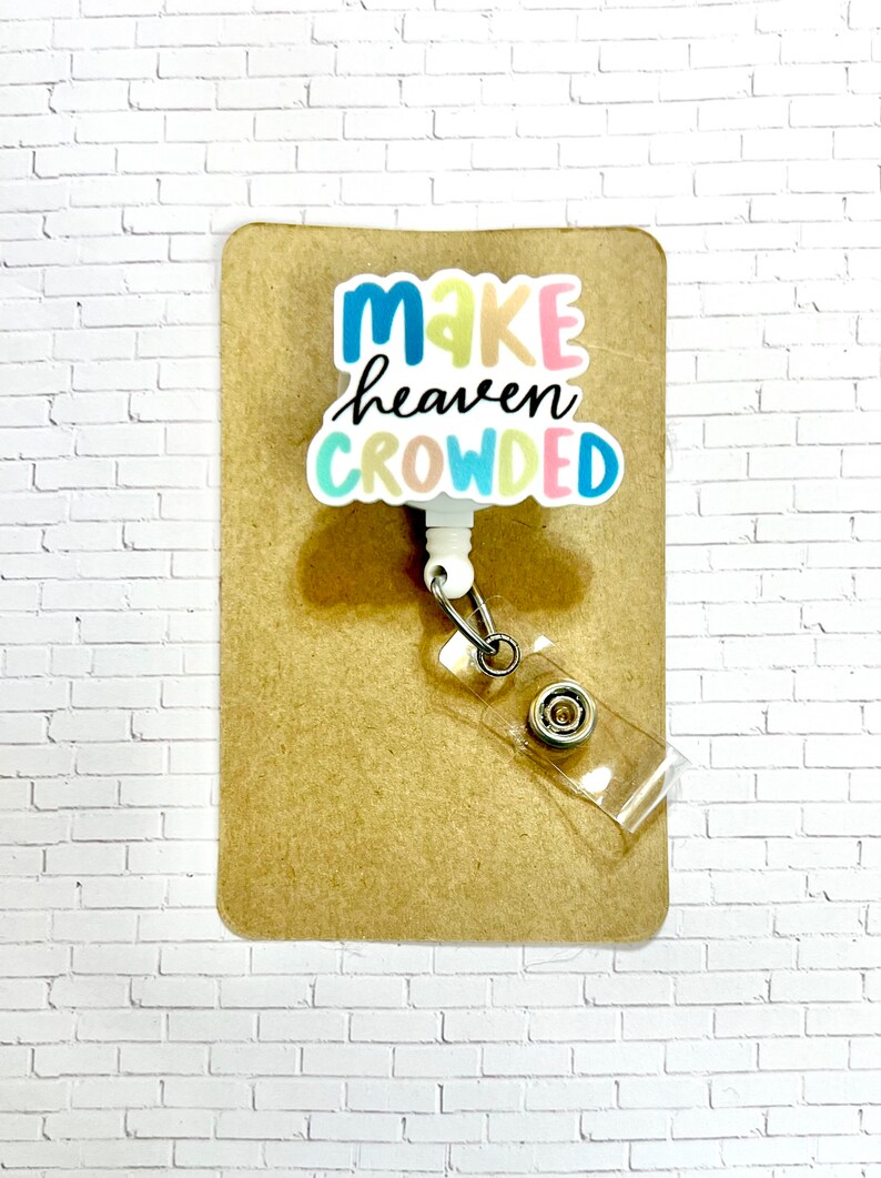May include: White retractable badge holder with a colorful "Make heaven crowded" graphic. The holder is on a brown background.