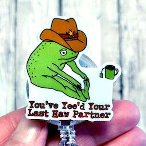 May include: A green frog wearing a brown cowboy hat with a gold star badge, sitting on a white background. The text "You've Yee'd Your Last Haw Partner" is displayed. A green teacup is also shown.