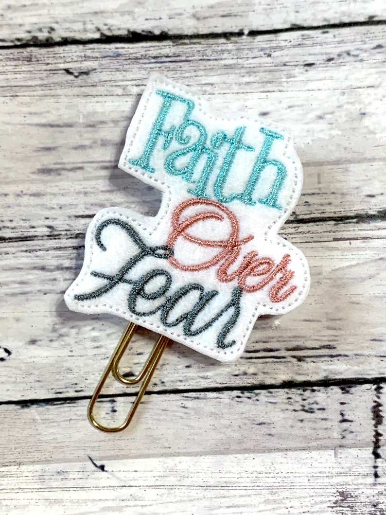 May include: White felt paper clip with the words "Faith Over Fear" embroidered in teal, pink, and gray thread.