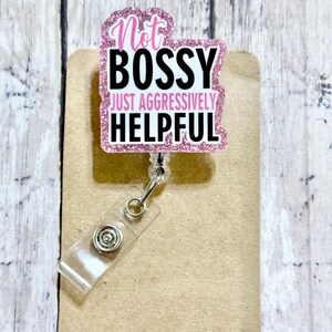 May include: A pink glitter badge reel with the text "Not Bossy Just Aggressively Helpful" in black and pink. The badge reel has a clear plastic strap and a metal clip.