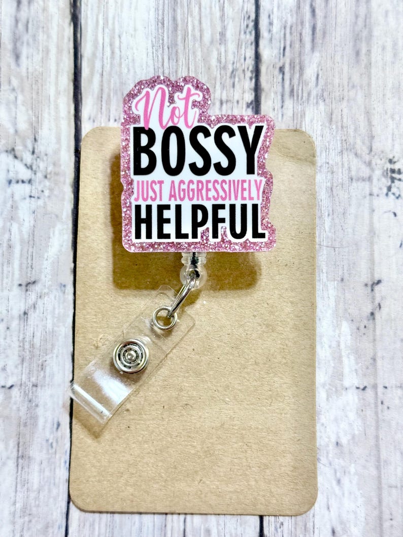 May include: A pink glitter badge reel with the text "Not Bossy Just Aggressively Helpful" in black and pink. The badge reel has a clear plastic strap and a metal clip.