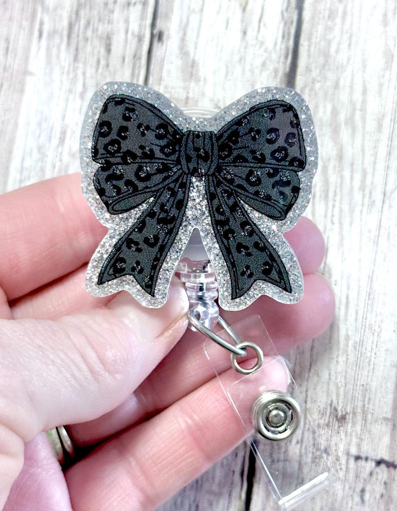 Puede incluir: Un portatarjetas en forma de lazo negro con un dise&ntilde;o de estampado de leopardo. El lazo est&aacute; delineado con purpurina plateada y tiene un cord&oacute;n retr&aacute;ctil de pl&aacute;stico transparente y un clip met&aacute;lico. El portatarjetas est&aacute; sostenido por una mano.