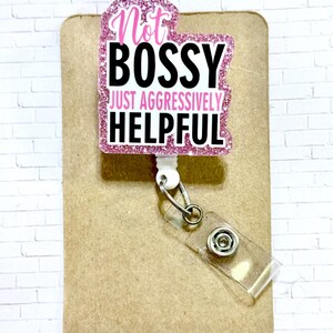May include: A pink glitter badge reel with the text "Not BOSSY JUST AGGRESSIVELY HELPFUL" in black and white. The badge reel is attached to a clear plastic holder and a metal clip. The background is a brown card.