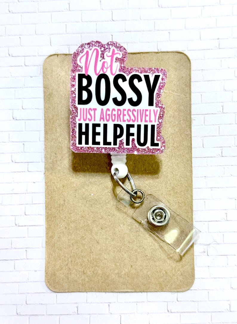 May include: A pink glitter badge reel with the text "Not BOSSY JUST AGGRESSIVELY HELPFUL" in black and white. The badge reel is attached to a clear plastic holder and a metal clip. The background is a brown card.