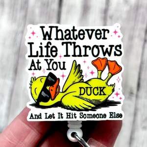 May include: A white badge reel with a cartoon duck wearing sunglasses. The badge has the text "Whatever Life Throws At You, And Let It Hit Someone Else". The duck is yellow with orange feet.