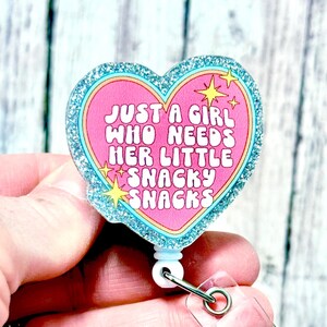 May include: A heart-shaped badge reel with a glittery silver border and the text "JUST A GIRL WHO NEEDS HER LITTLE SNACKY SNACKS" in white letters on a pink background. The reel has a white plastic base and a clear plastic clip.