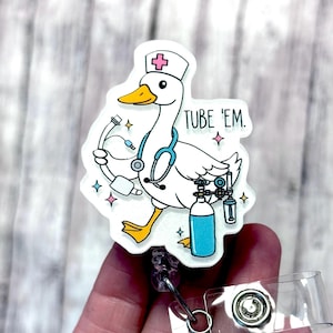 May include: A white badge reel featuring a cartoon goose wearing a nurse's cap and stethoscope, holding medical equipment. The text "TUBE 'EM" is printed on the badge. The design includes blue, pink, and yellow accents.