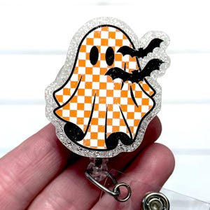 May include: A Halloween-themed badge reel featuring a smiling ghost with an orange and white checkered pattern. Two black bats are flying near the ghost. The badge reel has a clear, glittery border and a retractable cord.