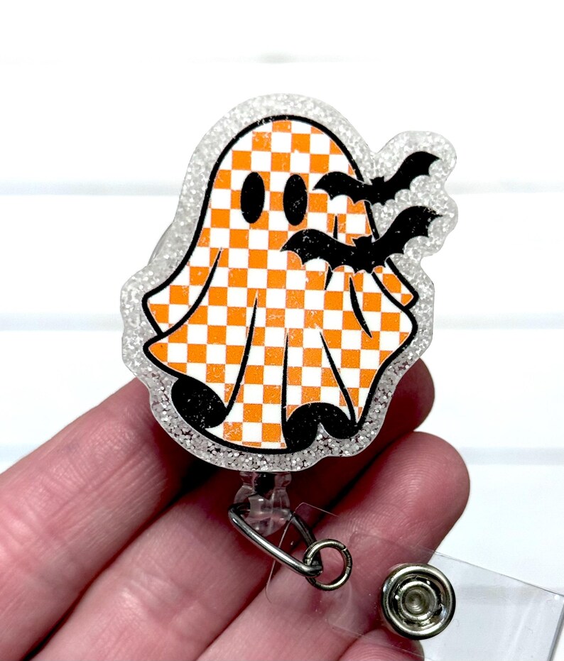 May include: A Halloween-themed badge reel featuring a smiling ghost with an orange and white checkered pattern. Two black bats are flying near the ghost. The badge reel has a clear, glittery border and a retractable cord.