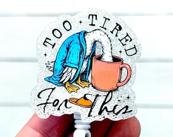 May include: A badge reel with a cartoon duck wearing a blue robe, with its head in an orange mug. The text reads "Too Tired For This". The badge reel is white with a retractable cord.