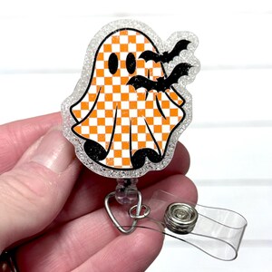May include: A Halloween-themed badge reel featuring a ghost with an orange and white checkered pattern and two black bats. The badge reel has a clear plastic clip and a retractable cord. The ghost is outlined with glitter.
