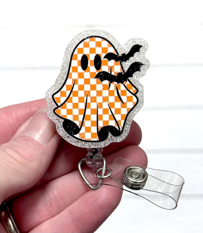 May include: A Halloween-themed badge reel featuring a ghost with an orange and white checkered pattern and two black bats. The badge reel has a clear plastic clip and a retractable cord. The ghost is outlined with glitter.