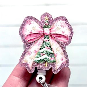 May include: A Christmas-themed badge reel featuring a pink glittery bow and a decorated Christmas tree. The bow has a snowflake and floral pattern. The tree is green with pink ornaments and a gold star.