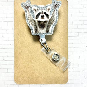 May include: A white retractable badge holder with a glitter-covered raccoon design. The raccoon is shown with its arms raised in the air.
