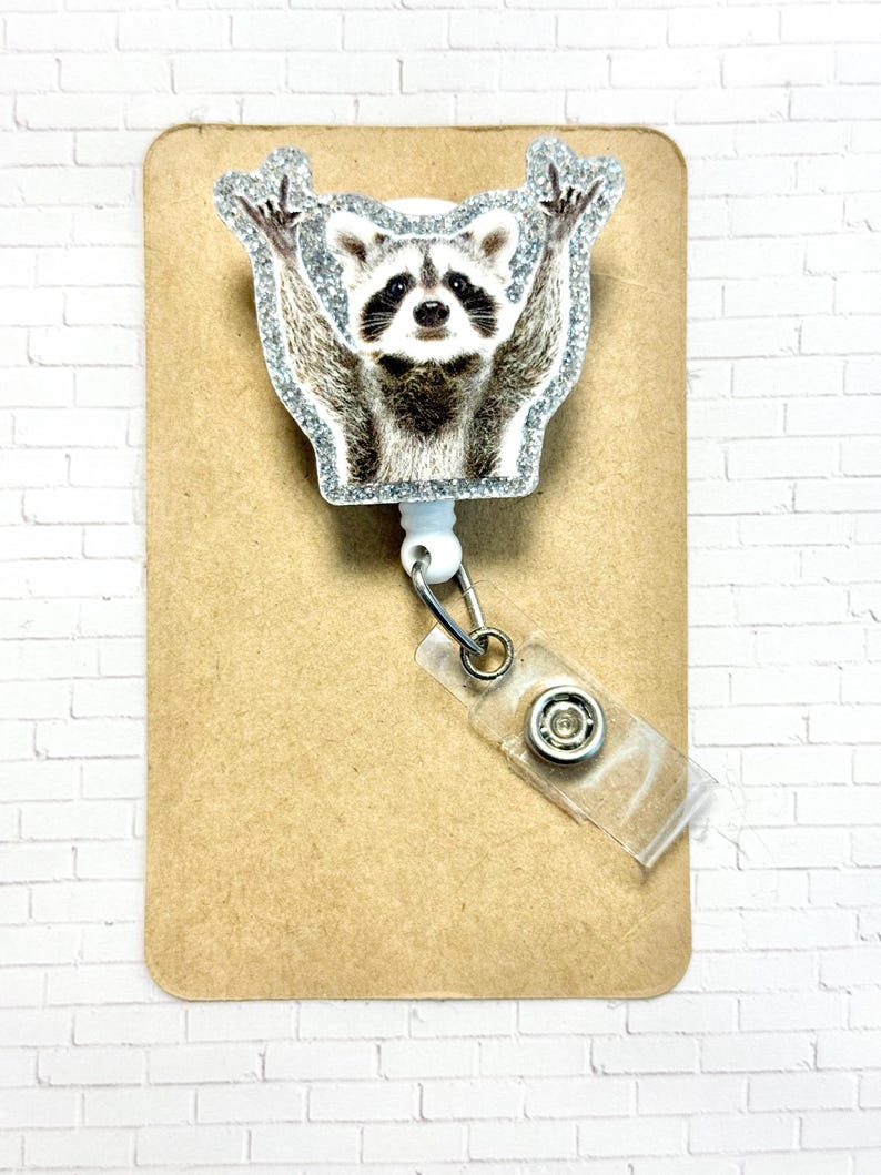 May include: A white retractable badge holder with a glitter-covered raccoon design. The raccoon is shown with its arms raised in the air.