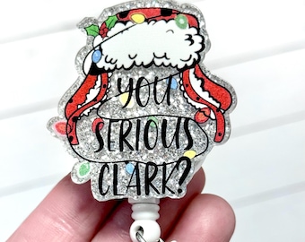 May include: A glittery badge reel with the phrase "YOU SERIOUS CLARK?" in black lettering. The badge features a Santa hat design with red and white accents, holly, and colorful Christmas lights.