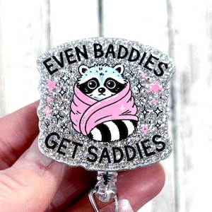 May include: A silver glitter badge reel with the text "EVEN BADDIES GET SADDIES". The badge features a cartoon raccoon wrapped in a pink blanket. Pink stars and silver sparkles surround the raccoon.