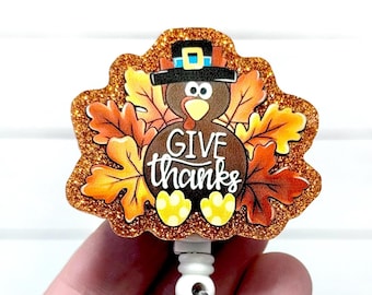 May include: Thanksgiving-themed badge reel featuring a turkey wearing a pilgrim hat, surrounded by autumn leaves. The text "GIVE thanks" is displayed in a cursive font. The badge reel has a glittery, orange border and a white retractable clip.