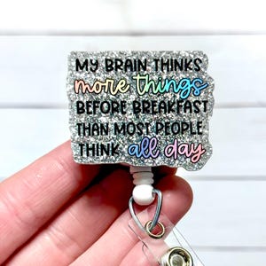 May include: A glittery badge reel with the text "MY BRAIN THINKS more things BEFORE BREAKFAST THAN MOST PEOPLE THINK all day" in colourful lettering. The badge reel has a clear plastic strap and a metal clip.