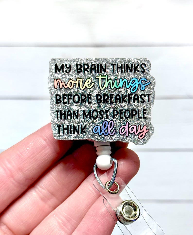 May include: A glittery badge reel with the text "MY BRAIN THINKS more things BEFORE BREAKFAST THAN MOST PEOPLE THINK all day" in colourful lettering. The badge reel has a clear plastic strap and a metal clip.