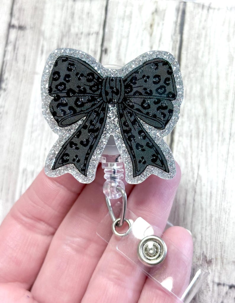 Puede incluir: Un carrete de insignia con un lazo negro con un dise&ntilde;o de estampado de leopardo. El lazo est&aacute; delineado con purpurina plateada. El carrete tiene una base de pl&aacute;stico transparente y un clip met&aacute;lico.