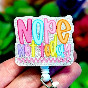 May include: A rectangular badge reel with the words "NOPE NOT TODAY" in colorful bubble letters. The letters are outlined in various colors, including pink, blue, orange, yellow, and purple, against a glittery background. The reel has a white plastic base and a metal clip.