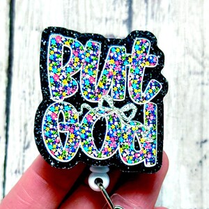 May include: A black glitter badge reel with the words "But Go Off" in a colorful floral pattern. The badge reel has a white retractable cord and a clear plastic clip.