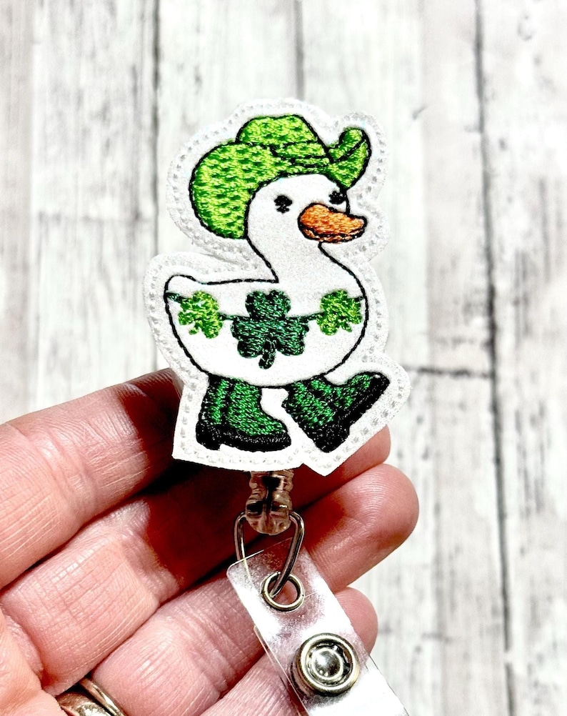 May include: White duck wearing a green hat and green boots with shamrocks around its neck. The duck is attached to a clear plastic retractable badge holder.