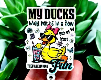 May include: A badge reel with a cartoon duck wearing sunglasses and a pink bow, holding a drink. The text reads "MY DUCKS may not be in a row But at least THEY ARE HAVING Fun."
