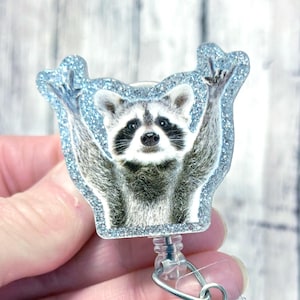 May include: A blue glitter badge reel with a raccoon wearing a blue glitter frame. The raccoon is holding its paws up in the air.