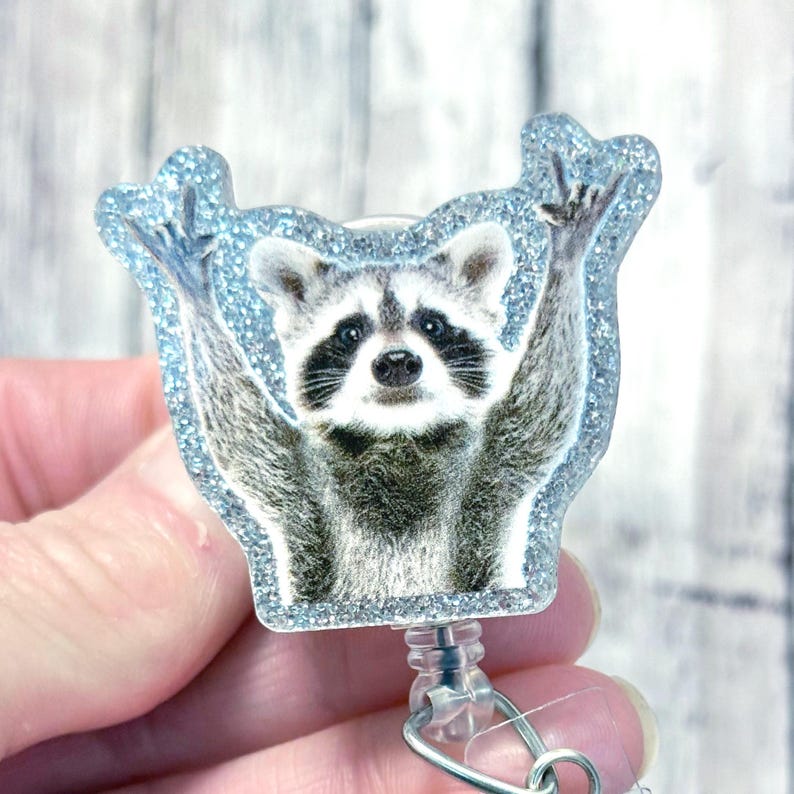 May include: A blue glitter badge reel with a raccoon wearing a blue glitter frame. The raccoon is holding its paws up in the air.