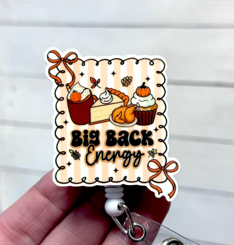 May include: A square badge reel with a Thanksgiving theme. The design features a variety of fall foods, including a pumpkin spice latte, pie, and a cupcake. The text "Big Back Energy" is in the center. The reel has a white retractable cord and a metal clip.