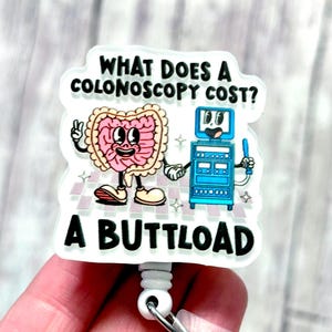 May include: A white badge reel with a cartoon illustration. The image features a smiling pink intestine character and a blue medical machine. Text on the badge reads "WHAT DOES A COLONOSCOPY COST? A BUTTLOAD."