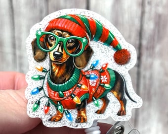 May include: A festive badge reel featuring a dachshund wearing a red and green Christmas sweater, hat, and glasses. The dog is adorned with colorful Christmas lights. The design is on a white background with a retractable reel.