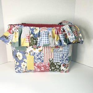 May include: A quilted tote bag featuring a patchwork design of various floral, gingham, and striped fabric patterns in shades of blue, yellow, red, and green. The bag includes a ruffled top edge and a single fabric strap with a matching pattern.