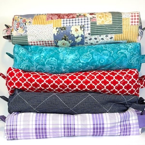 May include: A stack of six colorful bolster pillows. The top pillow has a patchwork design with floral and geometric patterns. The other pillows are turquoise, red with a white quatrefoil pattern, navy blue with white polka dots, and purple plaid.