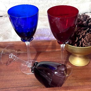 May include: Three colored wine glasses, two upright and one on its side, with a gold-colored bowl of pinecones in the background. The glasses are blue, red, and purple. The glasses have a bulbous design at the base of the stem.