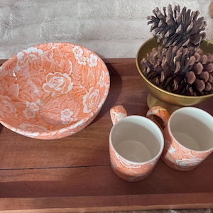 Peut inclure: Un plateau en bois présente un bol pêche et deux tasses assorties, tous ornés d'un motif floral blanc. Un bol doré avec des pommes de pin est posé sur le plateau. Le plateau a des poignées.
