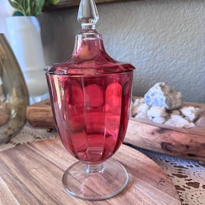 May include: A cranberry red glass candy dish with a matching lid featuring a pointed finial. The vessel has a vertical paneled design and sits on a clear circular glass pedestal base, showcasing a translucent finish and a polished surface.