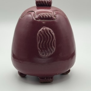 May include: A maroon-colored, egg-shaped ceramic container with a lid and three small feet. The container has a textured design on the lid and a wavy pattern on the front. The container is likely used for storage or decoration.