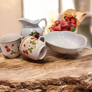 Könnte beinhalten: Eine Sammlung weißer Keramikgeschirr mit Erdbeermuster, darunter ein Krug, zwei Tassen und eine Schüssel. Frische Erdbeeren quellen aus einer braunen Papiertüte und setzen Farbakzente auf der rustikalen Holzoberfläche.