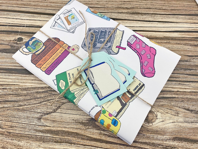 May include: A white gift wrap with a brown twine bow. The wrap features illustrations of books, socks, and mugs with book titles like "The Hobbit", "The Fellowship of the Ring", "The Two Towers", "The Return of the King", "Jane Eyre", "The Judes", "Harder We", and "Tlogers".