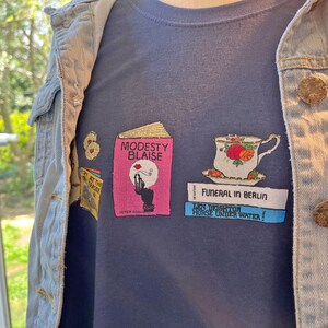 May include: A dark blue t-shirt with a graphic design featuring a pink book titled "Modesty Blaise", a teacup with flowers, and three books with the titles "Don't at the Seaside", "Funeral in Berlin", and "Len Deighton Horse Under Water!"