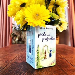 Op de afbeelding: Een keramische vaas beschilderd met de covers van de romans van Jane Austen, "Pride and Prejudice" en "Emma". De vaas is gevuld met gele bloemen.